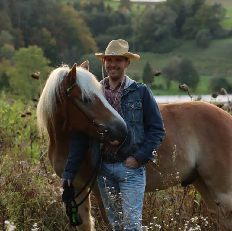 Ben, einen Cowboyhut tragend, steht auf einer Wiese und hält einen Haflinger am Zügel.